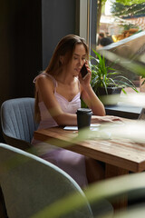 beautiful young asian woman sitting at coffee shop table surrounded by plants, working on laptop. beautiful lady calling by smartphone. modern online education, distant work, remote job, freelance