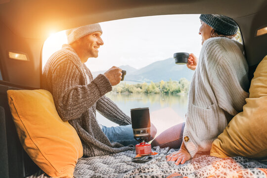 Car trunk view of chatting couple dressed warm knitted clothes enjoying gas stove prepared coffee and mountain lake view. Cozy early autumn couple auto traveling concept image.