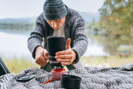 A man dressed in warm knitted clothes portrait starting a gas burner stove in the cozy car trunk with a beautiful mountain lake view. Warm early autumn auto traveling concept image.