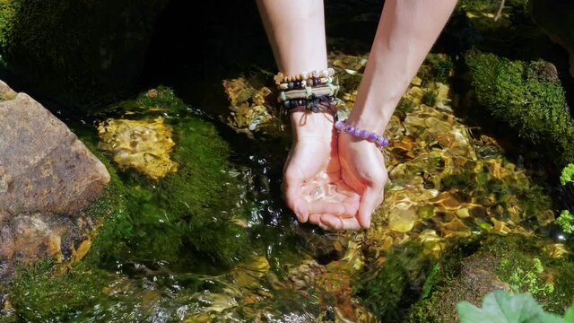 Female hands with boho bracelets scoop up clear water from stream to drink or wash. Current of clean spring flow over stones overgrown with moss. Girl in bijouterie quenches her thirst from river