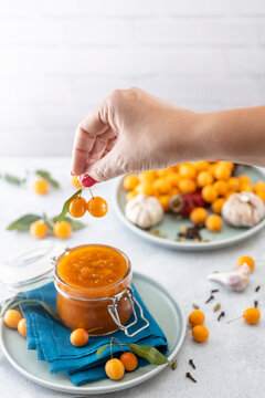 Woman hand holds yellow cherry plum over glass jar of homemade DIY natural canned yellow cherry plum sauce chutney with chilli or tkemali standing on white table