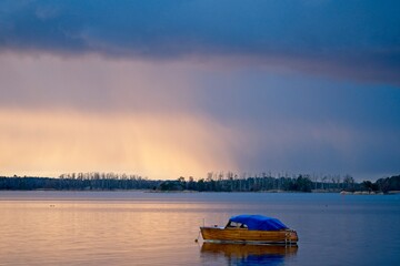 Small wooden boat with blue cover on lake. Burning agitated stormy skies in the background.