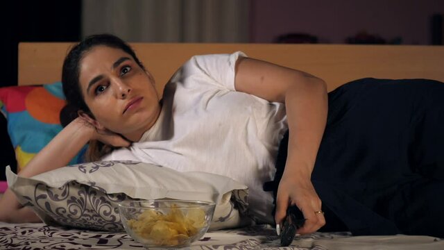An attractive lady with black hair eating chips / wafers while watching television - Binge eating. A pretty female surfing TV channel using a remote control - electronic device  modern lifestyle  l...