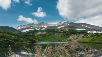 The emerald water of the lake is calm. A mountain range against a background of blue sky and clouds. There are areas of snow on the slopes. In the foreground - spikelets of grass. Kamchatka