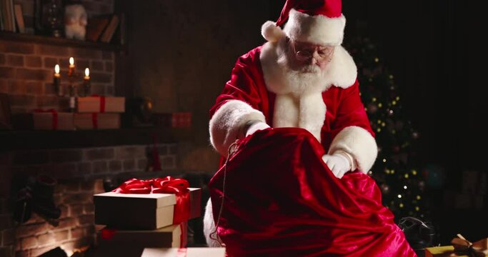 Santa Claus putting toys and taking heavy red bag with gifts close-up, carrying big sack full of presents for children. Christmas atmosphere concept. Burning fireplace and green tree on background.