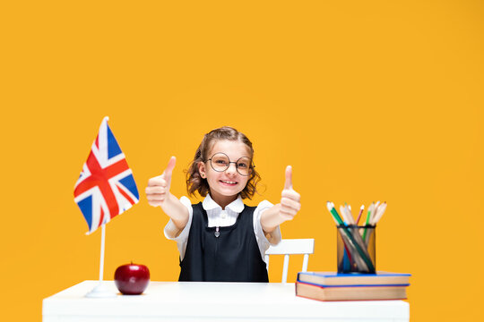 Happy caucasian schoolgirl sitting at the desk showing thumbs up. English lesson Great Britain flag
