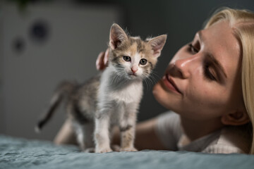 A woman's face bent over the bed with a small gray-white kitten and strokes it. The development and growth of a baby cat. Love for pets. Close-up, blurred background.