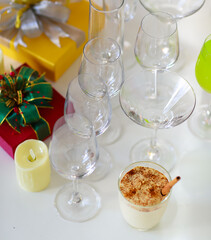 Tasty delicious milk cream eggnog drink with gingerbread in glass on table in front empty tall wine glasses present gift boxes white chocolate candles green apple punch and decorated Christmas tree