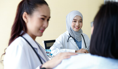 Close up shot of Muslim Arab Islam wears hijab in white lab coat uniform with stethoscope typing information in laptop computer while Asian female doctor talking with patient in blurred foreground