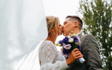 Portrait of newlyweds wedding kiss outdoors, groom in suit kissing bride in white dress with veil and bouquet, low angle view. Wedding ceremony, young husband and wife, marriage and love concept