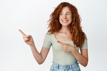 Happy young woman with natural long hair and freckles, pointing and looking left with pleased smile, showing advertisement beside, standing over white background