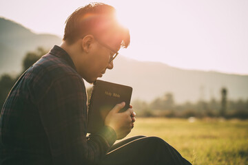 man praying on the holy bible in a field during beautiful sunset.male sitting with closed eyes with...