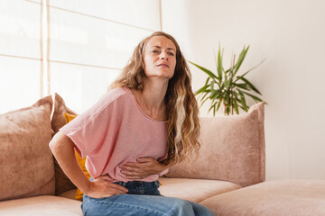 Young woman suffering from strong abdominal pain while sitting on sofa at home