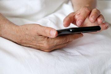 Elderly woman with smartphone in a bed, mobile phone in wrinkled female hands close up. Concept of online communication in retirement, sms, social media