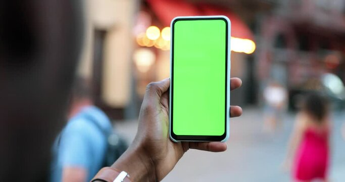 Close up of the hand of multiracial woman holding vertical black smartphone with green screen and tapping on it on the street of the city. Chroma key concept