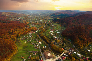 Aerial view of a village rural area with small houses between autumn mountain hills covered with yellow and green spruce forest.