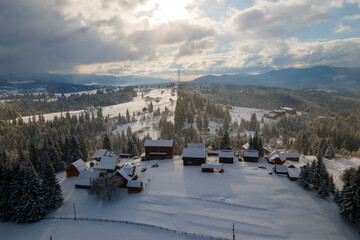 Aerial winter landscape with small village houses between snow covered forest in cold mountains in the evening.