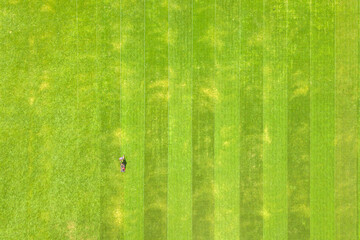 Aerial view of small figure of man worker trimming green grass with mowing mashine on football stadium field in summer. - obrazy, fototapety, plakaty