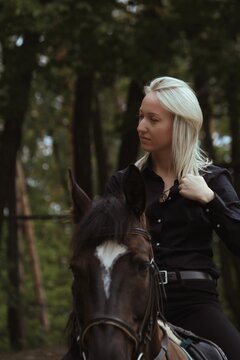 A Blonde Girl In Profile Riding A Black Horse Against A Green Forest. A Girl Is Riding A Brown Horse On A Autumn Day.