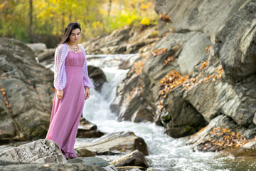 Young pretty woman in long fashionable dress standing near small mountain river with fast moving water.