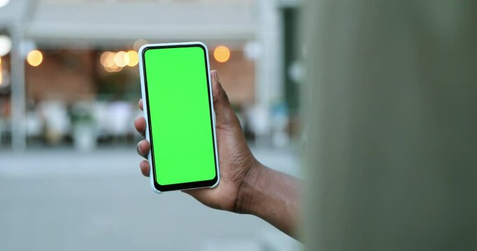 Male hands holding smartphone with green screen. Guy using mobile phone while walking in the autumn street. Back view shot. Chroma key, close up man hand holding phone with vertical green screen
