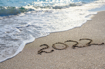 Inscription 2022 on a sandy beach in the afternoon. In the background, a blue sea wave washing the shore. Selective focus for 2022. Concept for the new 2022.