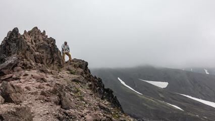 A man with a trekking pole stands on top of a cliff. Cracked stones on steep slopes devoid of vegetation. In the background-mountain slopes with patches of snow. The peaks are shrouded in thick fog.