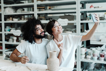A man and woman in love take a selfie in a pottery workshop, sitting at a table with a clay pot and brushes for painting. Dating in a hobby studio, spending leisure time
