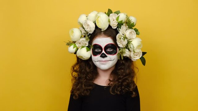 Close up portrait of bright little girl kid with Halloween makeup mask wears black outfit and flowers wreath, keep eyes closed open smile isolated over yellow background. Celebration party concept