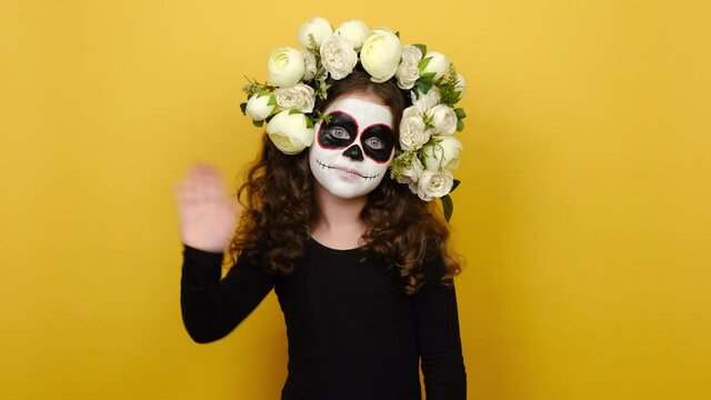 Portrait of cute happy girl kid with halloween or day of death makeup, dressed in black outfit and flowers wreath, waving her hand in hello gesture standing isolated over yellow studio background wall