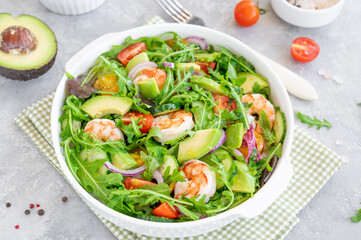 Delicious fresh salad with prawns, tomatoes, avocado, cucumber and onions in a bowl on a gray concrete background. Healthy dish. Top view, copy space.
