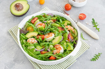 Delicious fresh salad with prawns, tomatoes, avocado, cucumber and onions in a bowl on a gray concrete background. Healthy dish. Top view, copy space.