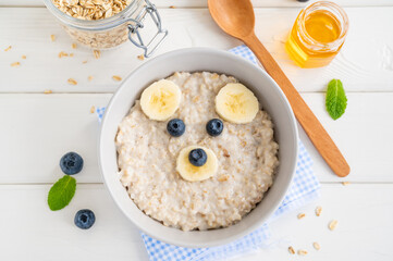 Funny bowl with oat porridge with bear faces made of fruits and berries on a white wooden background. Food for kids idea, top view, copy space
