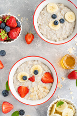 Funny bowls with oat porridge with owl and bear faces made of fruits and berries on a gray concrete...