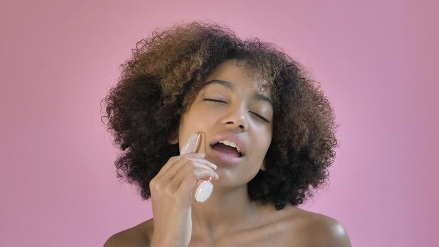 Relaxed black young woman with short curly hair massages neck with special jade massager roller against pink background closeup