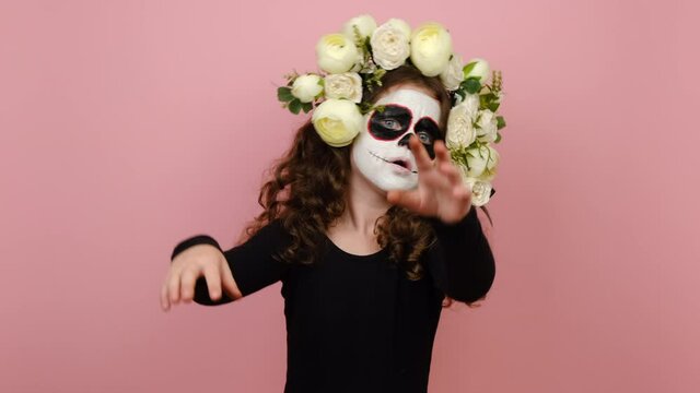 Spooky little girl with Halloween makeup mask wears black outfit and flowers wreath hands raised, scares looking at camera, isolated on color pink background. Mexican culture, Day of Death concept