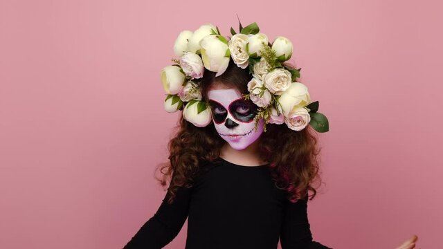 Spooky small girl child with sugar skull makeup, wears flowers wreath, scaring you by hands, saying boo, posing isolated on pink studio background wall. Day of Dead and Happy Halloween concept