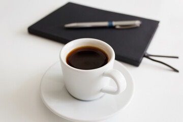 The concept of a workplace with coffee in a neat white mug on a saucer and a notebook with a pen on a modern white office desk. Break for a delicious and hot coffee. Minimalism. Side view
