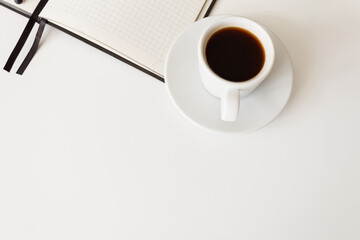 The concept of a workplace with coffee in a neat white mug on a saucer and an open notebook on a modern white office desk. Break for a delicious and hot coffee. Minimalism. Side view with copy space