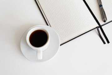 The concept of a workplace with coffee in a white mug on a saucer and an open notebook with a pen on a modern white table. Minimalism. Side view from above with a place to copy. Coffee break. Close-up