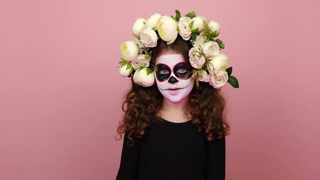 Little girl child with Halloween makeup mask, wears flowers wreath says Boo laughs, spooky looking at camera, posing isolated over pink color background in studio. Happy Halloween and Day of Death