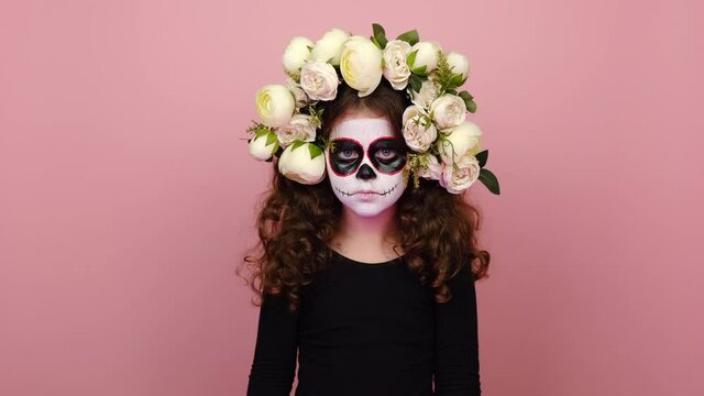 Serious severe girl kid with Halloween makeup mask wears black outfit and flowers wreath, say no hold palm folded crossed hands in stop gesture, isolated on yellow background. Day of Death concept