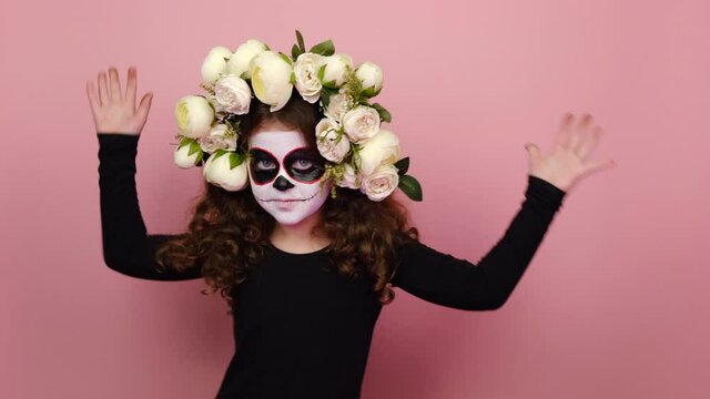 Scary little girl kid with Halloween makeup mask wears black outfit and flowers wreath dance gesticulating hands fool around have fun enjoy celebrate isolated on pink studio background. Day of Dead