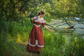 A young woman in a 19th century dress by the river. Girl with a book in his hand, summer day. The photo