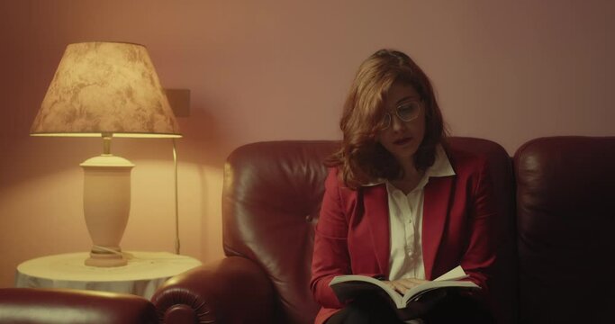 Giovane bella ragazza italiana con giacca rossa legge il libro seduta sul divano nella stanza di casa