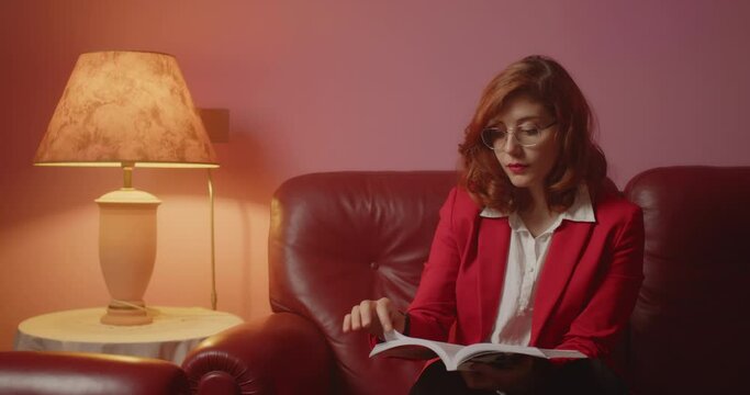 Giovane bella ragazza italiana con giacca rossa legge il libro seduta sul divano nella stanza di casa