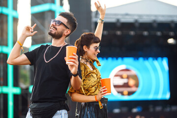 Stylish couple enjoying a performance at a festival together. Young people drinking beer and having fun at music festival at the beach. Youth, party, vacation concept.