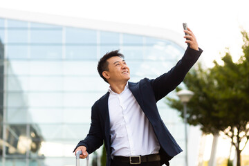 Asian tourist businessman arrived at a business conference takes photos near the airport and communicates by video with colleagues using a smartphone