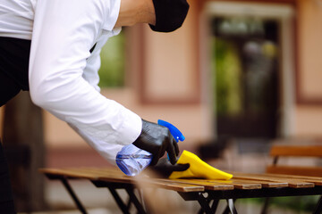 Disinfecting to prevent COVID-19. Waiter cleaning the table with Disinfectant Spray in a restaurant wearing protective medical mask and gloves.