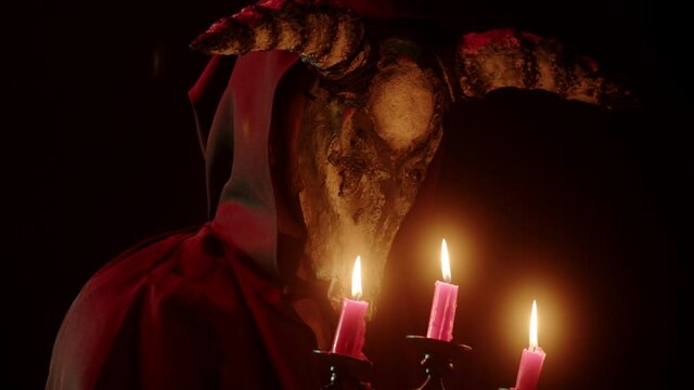 Devil with cow skull head holding burning candles. Demon with horns conjuring close-up. Evil imp wearing black robe with hood using witchcraft in darkness. Halloween and full moon.